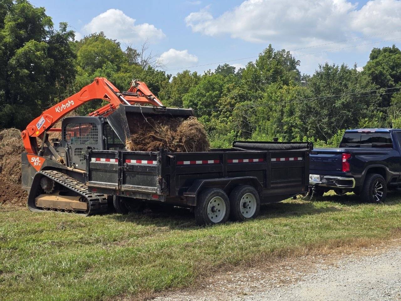 Orange track loader beside a black dump trailer and pickup truck loaded with cleared debris