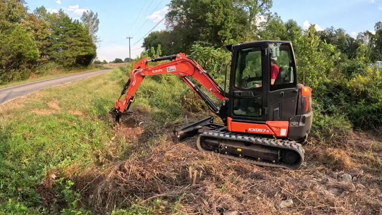 Orange mini excavator digging a trench in a grassy area near the woods for a culvert