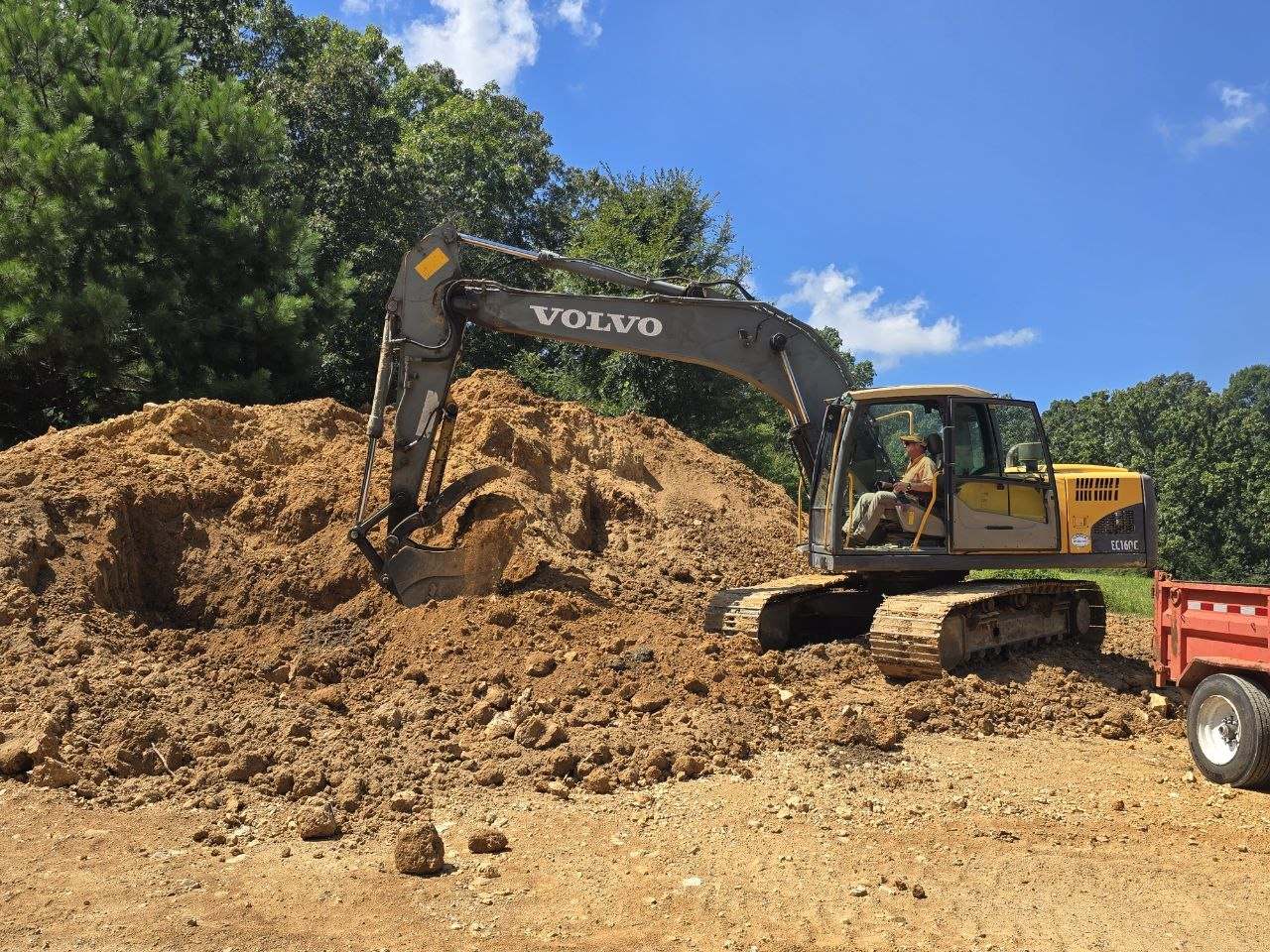 Large excavator digging into a big pile of dark clay under a blue sky