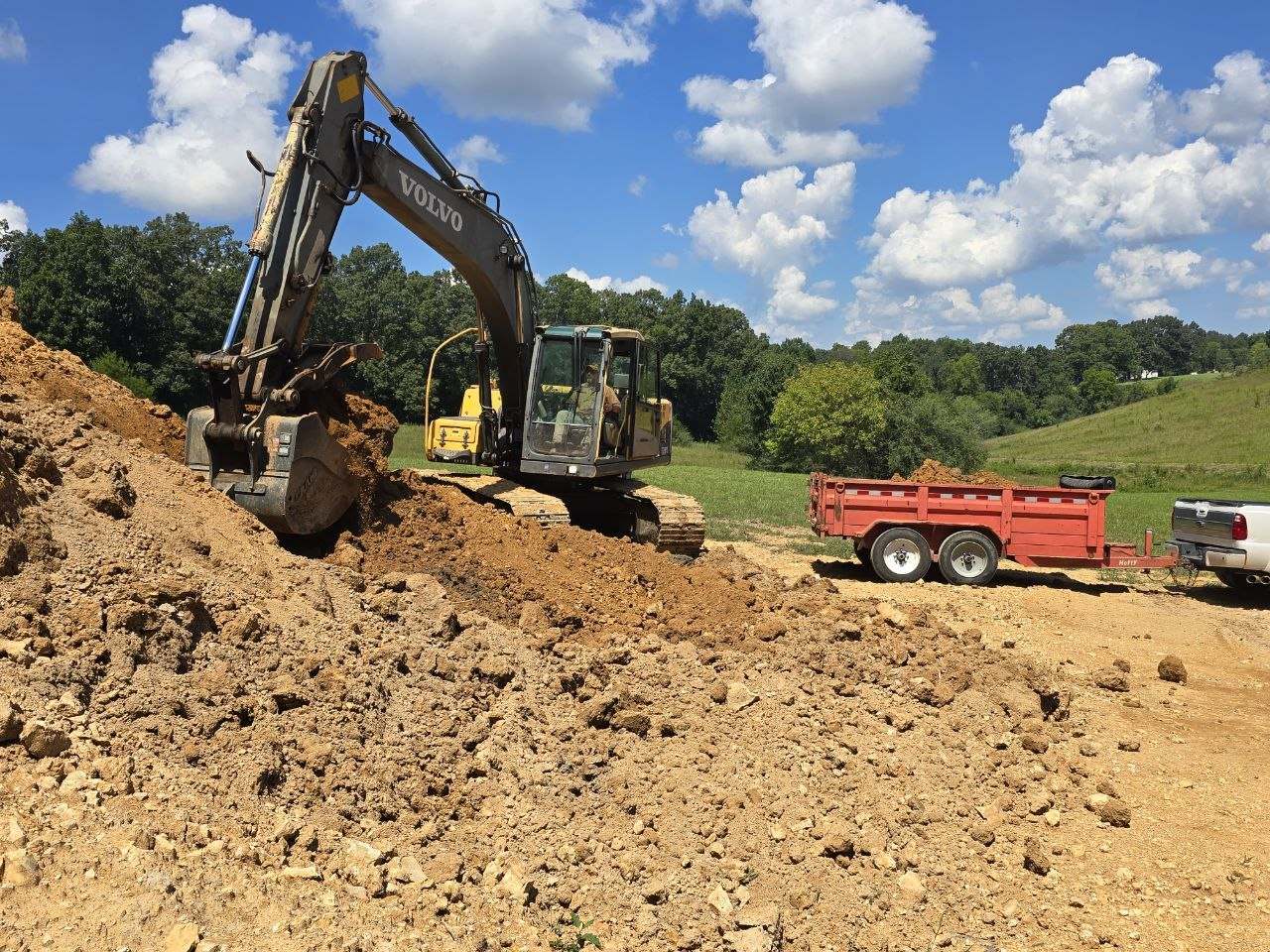 Excavator on a dirt slope continuing to load a red dump trailer