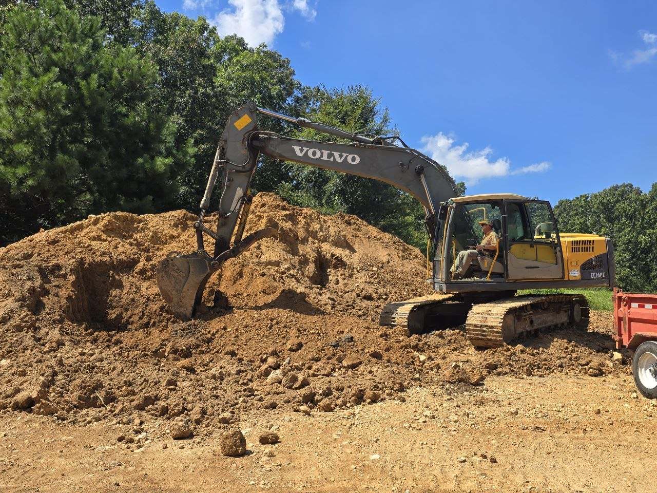 Excavator working a large soil mound with trees behind it
