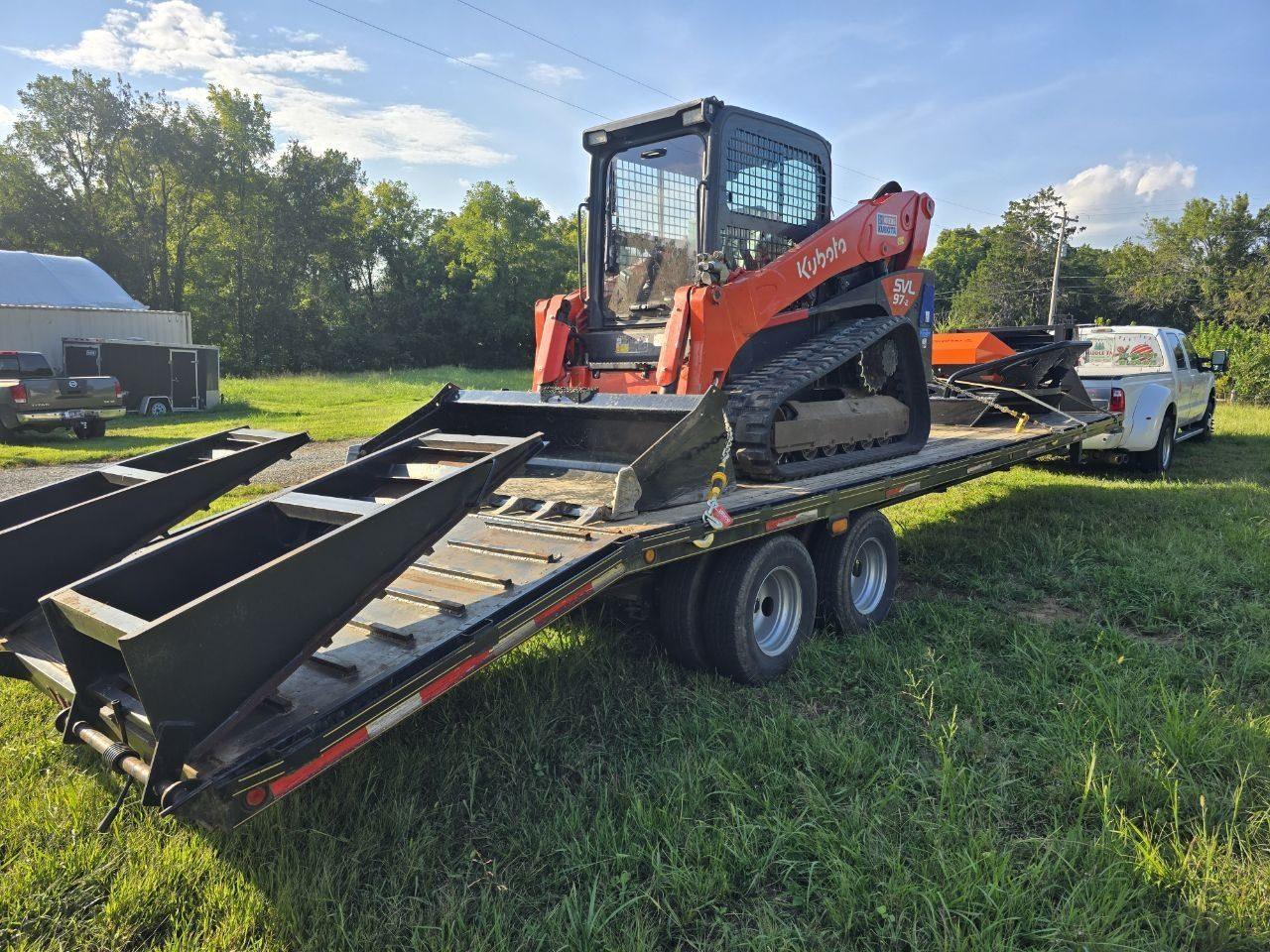Orange Kubota track loader strapped to a black flatbed trailer behind a white pickup truck, next to a white barn