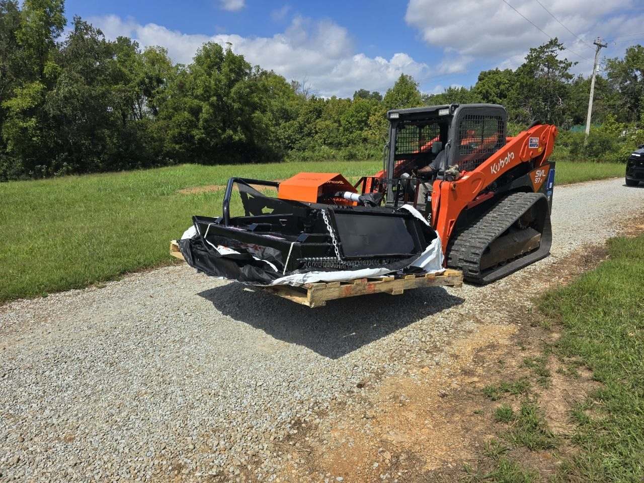 Front view of an orange Kubota track loader with a black brush hog attachment resting on a wooden pallet on a gravel drive