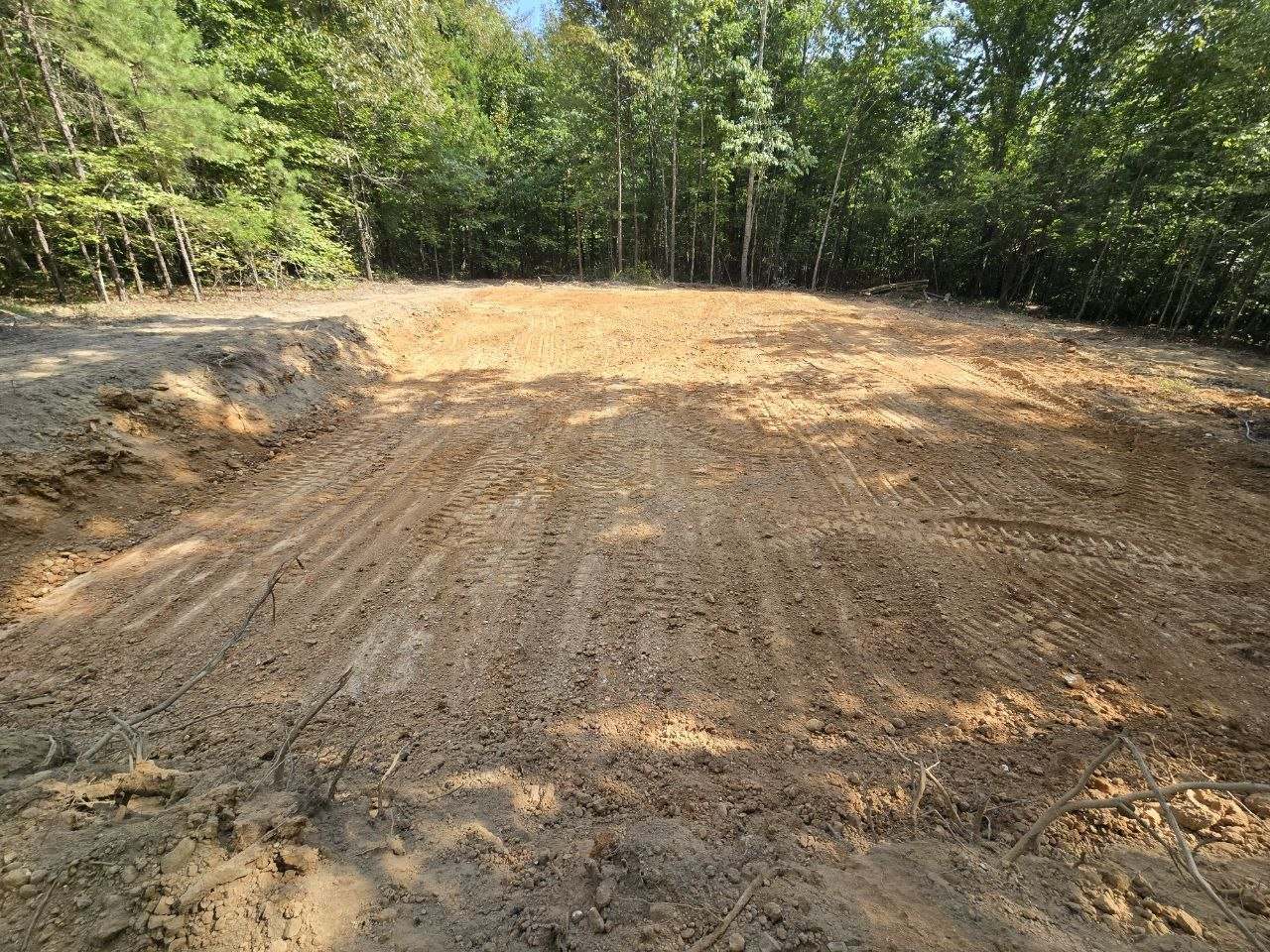 Large rectangular leveled pad of light brown soil with parallel track marks, bordered by woods on three sides