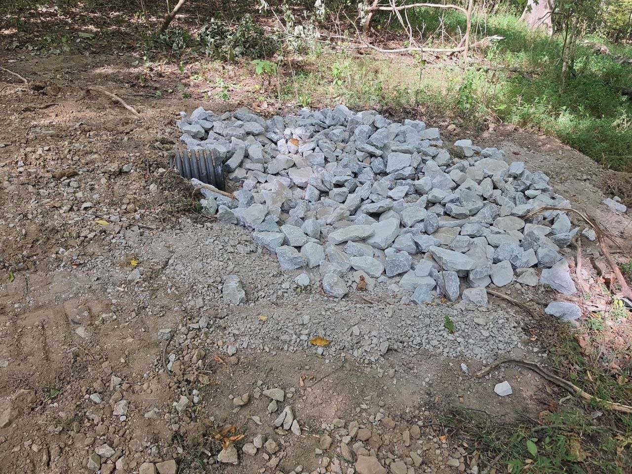Black corrugated culvert pipe outlet emerging from the ground, surrounded by a bed of large gray rip rap stone