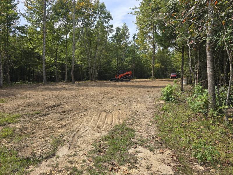 Large flat lot cleared down to bare dirt with fresh track marks, a skid steer at the far end and woods behind