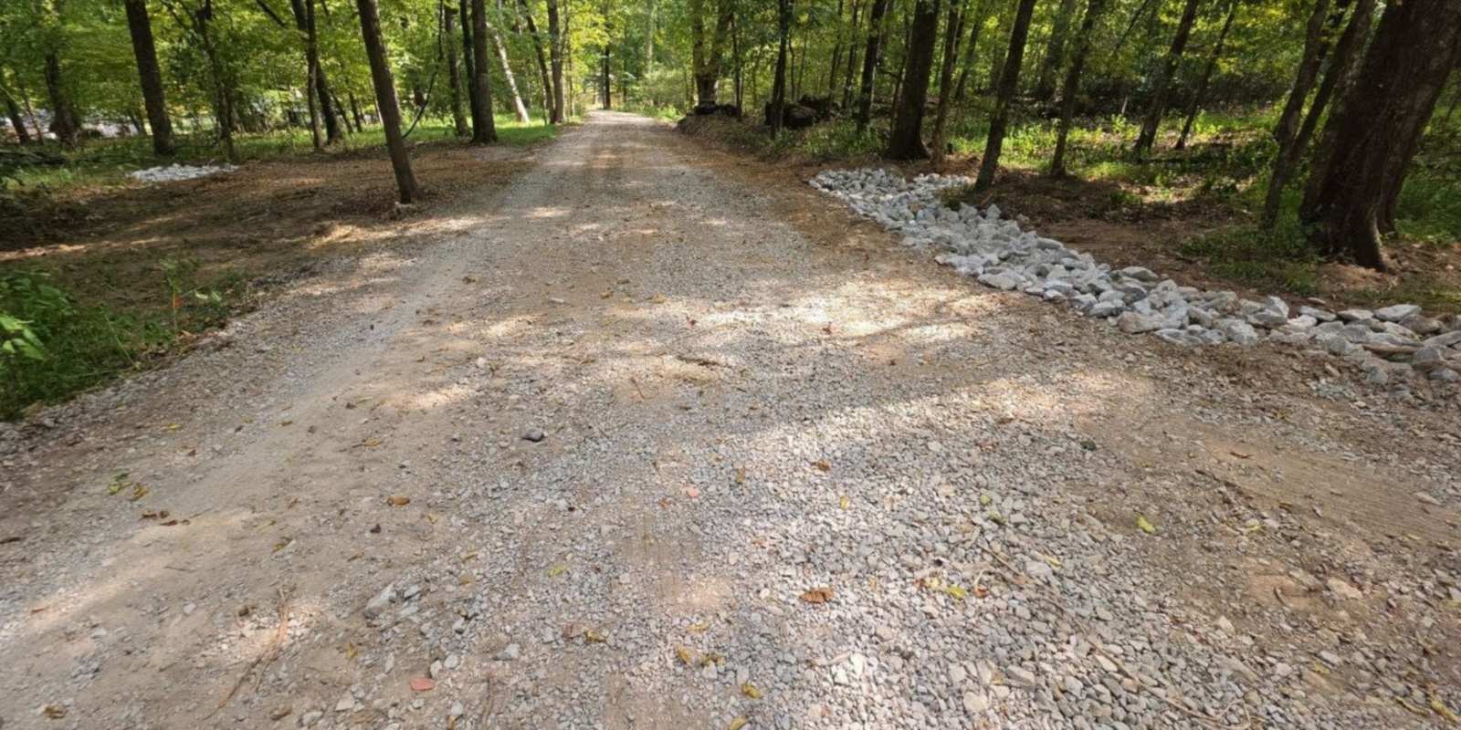 Finished gravel driveway running straight through the woods, with drainage rock along the edge