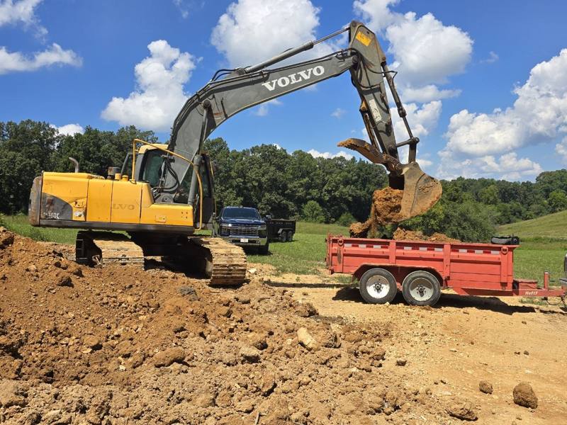 Excavator loading dirt into a red dump trailer hitched to a pickup truck, with trees in the background