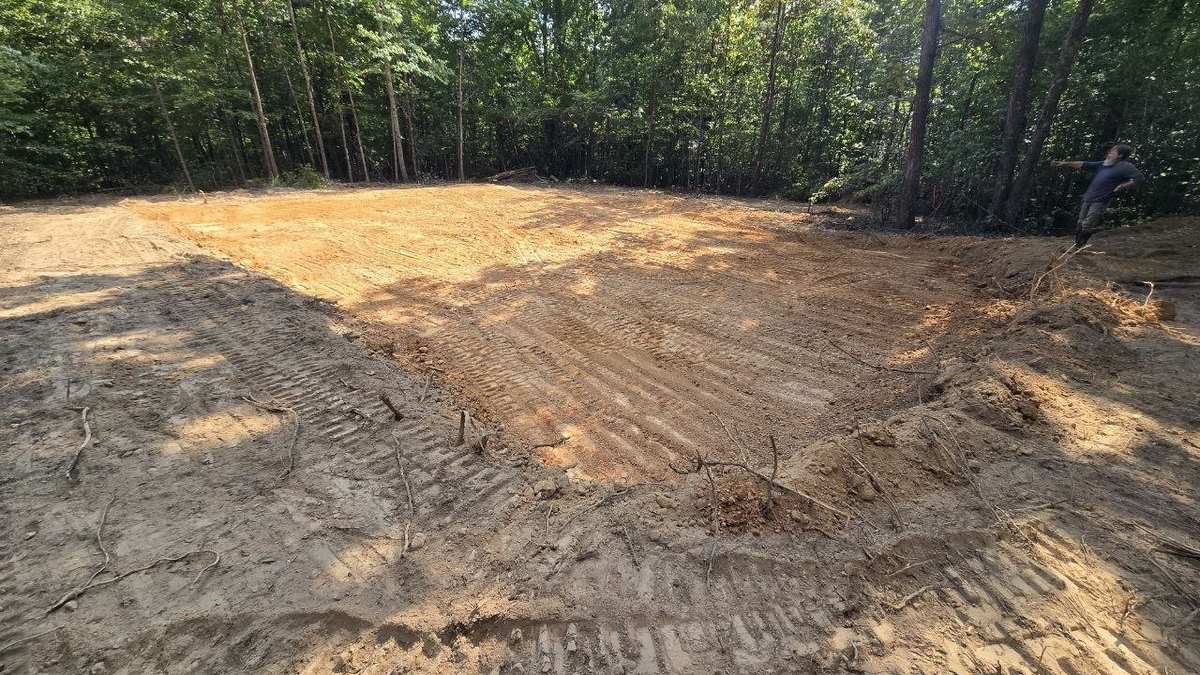 Freshly graded rectangular building pad with machine track marks, surrounded by woods
