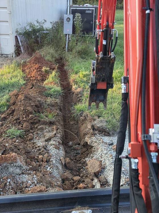 Narrow freshly dug trench running alongside a white building, with the orange excavator arm and a spoil pile beside it